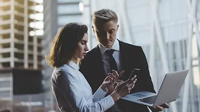 Two colleagues dressed in business attire outside interacting with a phone while the other holds a laptop.